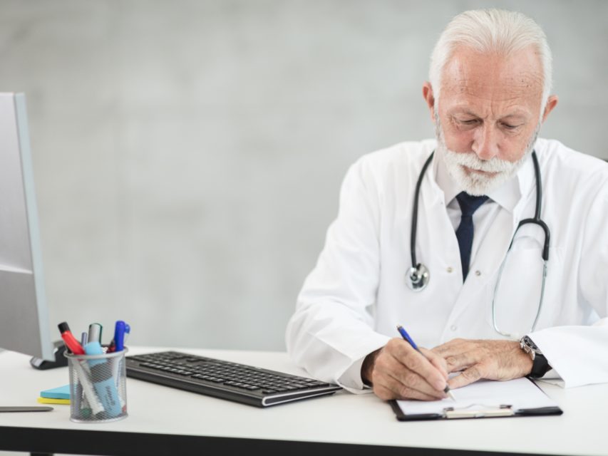 A physician at a desk, writing notes by hand next to a computer